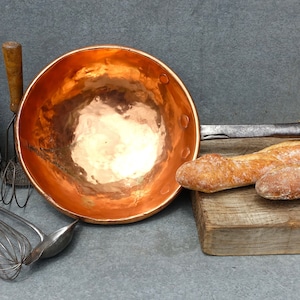 Puede incluir: Una olla grande de cobre con un mango de madera, tres panes, una botella de vino, un batidor, un colador de alambre y un cucharón sobre una superficie de madera.