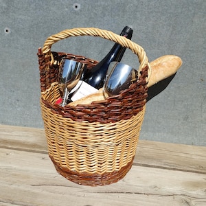Op de afbeelding: Een geweven picknickmand met een gevlochten handvat, met een fles wijn, twee roestvrijstalen wijnglazen en een brood. De mand is bruin en beige en staat op een houten oppervlak.