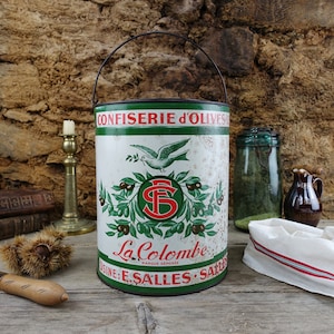 May include: A vintage metal container for olives, featuring a green and white design with a dove and the text "Confiserie d'Olives La Colombe Marque Deposee Usine: E. Salles - Salles".