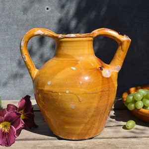 May include: A large, yellow earthenware pitcher with two handles. The pitcher has a rustic, antique look and is made of glazed ceramic.
