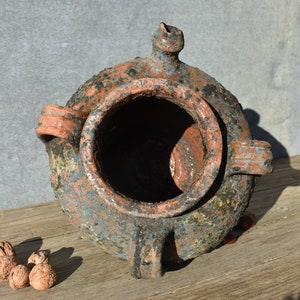 Pu&ograve; includere: Un grande vaso rustico in terracotta con un'ampia apertura e tre manici. Il vaso &egrave; ricoperto da uno strato di terra e muschio, che gli conferisce un aspetto invecchiato e consumato.
