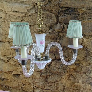 May include: A white and pink glass chandelier with three light bulbs and green lampshades. The chandelier has a twisted glass design and is hanging from a chain.