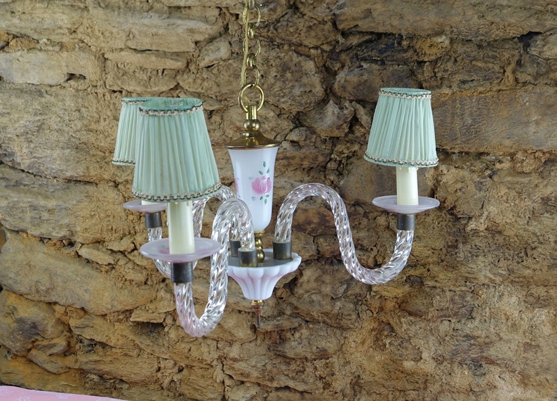 May include: A white and pink glass chandelier with three light bulbs and green lampshades. The chandelier has a twisted glass design and is hanging from a chain.