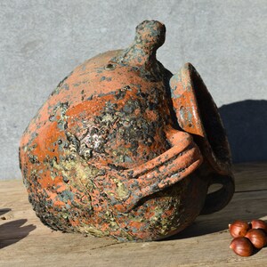 Pu&ograve; includere: Un grande vaso rustico in terracotta con una smaltatura screziata arancione, marrone e blu. Il vaso ha un'ampia apertura e un manico sul lato.