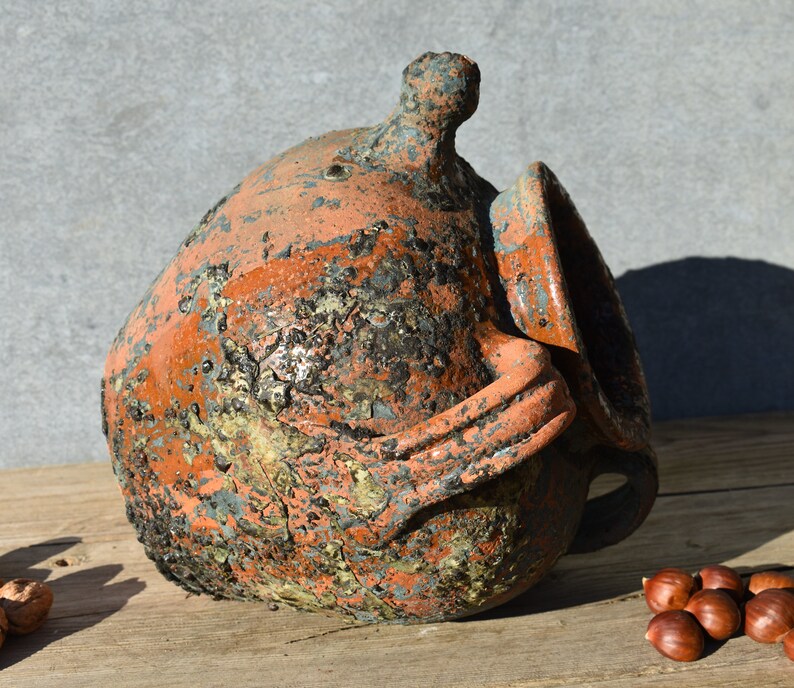 Pu&ograve; includere: Un grande vaso rustico in terracotta con una smaltatura screziata arancione, marrone e blu. Il vaso ha un'ampia apertura e un manico sul lato.