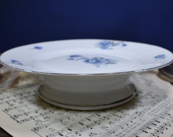 Op de afbeelding: Een witte porseleinen taartplateau met een zilveren rand en een blauw bloemenontwerp.