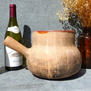 Puede incluir: Una olla grande de barro marrón con un asa de madera. La olla tiene un cuerpo ancho y redondo y una abertura estrecha. La olla está sobre una superficie gris junto a dos botellas de vino.