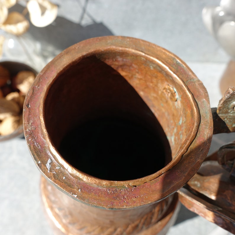 K&ouml;nnte beinhalten: Ein Kupferkrug mit einer breiten &Ouml;ffnung und einem Henkel. Der Krug hat ein abgenutztes, gealtertes Aussehen mit einer Patina aus Gr&uuml;n und Braun.