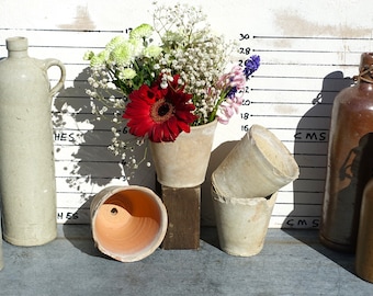 May include: Four terracotta flower pots, two with white paint, one with a red gerbera daisy, and one empty. The pots are arranged on a wooden surface with two glass bottles in the background.