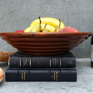 Könnte beinhalten: Eine Holzschale gefüllt mit gelben Bananen, grünen Birnen und roten Äpfeln. Die Schale steht auf zwei schwarzen Büchern mit goldenen Schriftzügen. Im Hintergrund stehen zwei Flaschen Wein.
