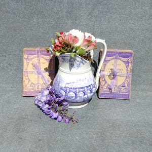 Puede incluir: Una jarra de porcelana azul y blanca con un diseño floral se encuentra sobre una superficie gris. La jarra está llena de flores rosas y blancas. Tres libros antiguos con cinta morada están apilados detrás de la jarra. Los libros se titulan "Classique Larousse" y "Bovet Oeuvres Choisies".