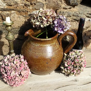 Op de afbeelding: Een rustig stilleven met een bruine keramische kruik met paarse en groene hortensia's, een messing kandelaar met een brandende kaars, een donkere wijnfles en roze hortensia's. De opstelling staat op een houten oppervlak.