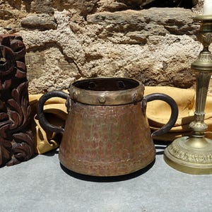 May include: A vintage copper mug with black handles and a brass rim is displayed beside a carved wooden panel and a brass candlestick. The mug has a hammered texture and a rustic look. The candlestick holds a white candle.