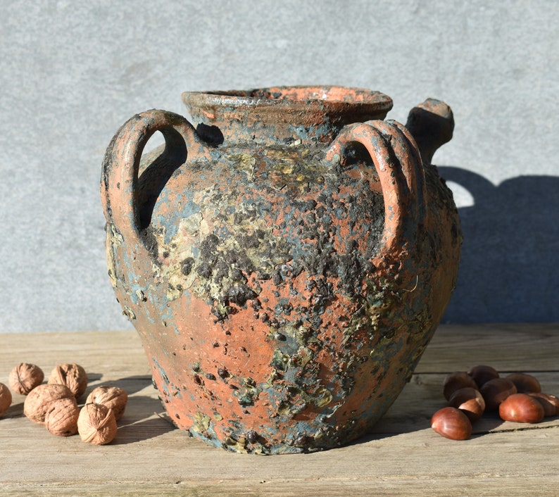 Pu&ograve; includere: Un grande vaso in terracotta rustico con due manici. Il vaso ha una finitura screziata e invecchiata con sfumature di marrone, blu e verde.