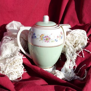 May include: A green and white ceramic teapot with floral designs and gold accents. The teapot is sitting on a red fabric surface with a white lace scarf.