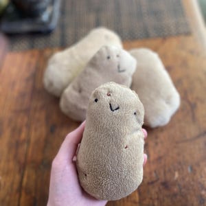 May include: A plush potato toy with a smiling face and small red accents is held in a hand. Several other similar plush potatoes are in the background on a wooden surface. The toys are beige and tan.