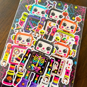 May include: A collection of die-cut stickers featuring colorful retro-style robots with Halloween-themed details like pumpkins, ghosts, bats, and skeletons. The robots are displayed on a black backing sheet decorated with a vibrant neon spiderweb pattern.