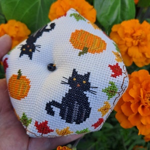 May include: A white, cross-stitched pincushion with black cat and pumpkin designs, plus autumn leaves. The pincushion has a black button and is set against orange flowers and green leaves.