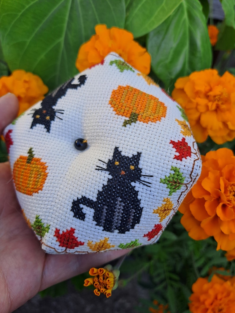 May include: A white, cross-stitched pincushion with black cat and pumpkin designs, plus autumn leaves. The pincushion has a black button and is set against orange flowers and green leaves.