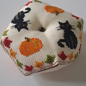 May include: A square, cross-stitched cushion with a black cat and orange pumpkins on an off-white background. The cushion is decorated with colourful autumn leaves around the edges and has a black button in the centre.