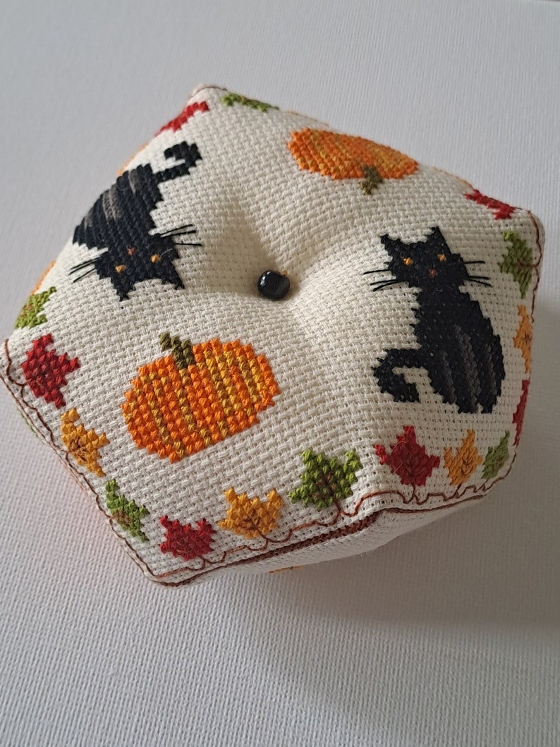 May include: A square, cross-stitched cushion with a black cat and orange pumpkins on an off-white background. The cushion is decorated with colourful autumn leaves around the edges and has a black button in the centre.