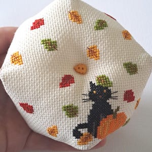 May include: A cream-coloured, diamond-shaped, cross-stitched pillow. It features a black cat on an orange pumpkin, surrounded by red, green, and gold autumn leaves. A small, round, tan button is sewn on the pillow. The design evokes a seasonal, autumnal theme.