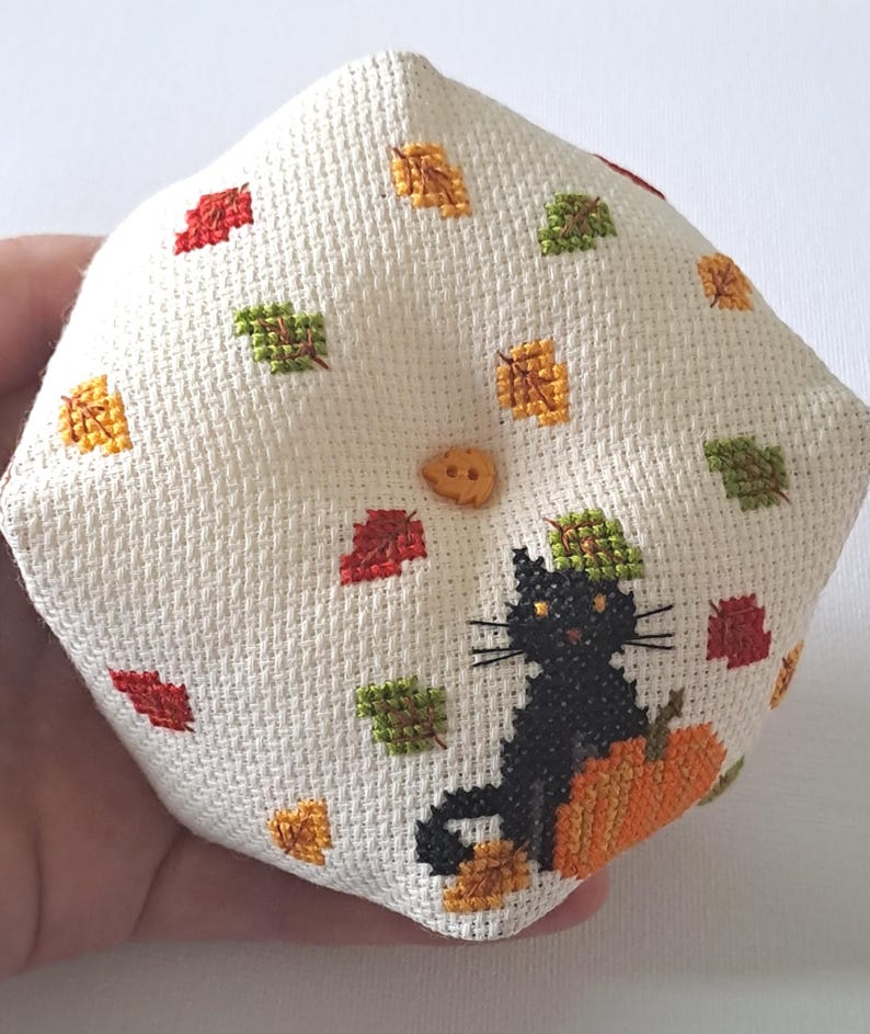 May include: A cream-coloured, diamond-shaped, cross-stitched pillow. It features a black cat on an orange pumpkin, surrounded by red, green, and gold autumn leaves. A small, round, tan button is sewn on the pillow. The design evokes a seasonal, autumnal theme.