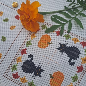 May include: Cross-stitch design with two black cats and two orange pumpkins, framed by autumn leaves. An orange flower and a gold sequin are placed on the white fabric. The design has a fall or Halloween theme.