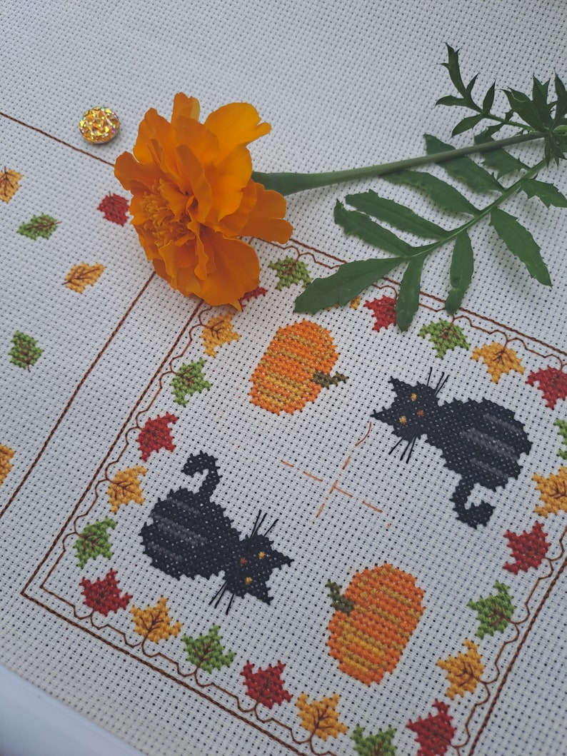 May include: Cross-stitch design with two black cats and two orange pumpkins, framed by autumn leaves. An orange flower and a gold sequin are placed on the white fabric. The design has a fall or Halloween theme.