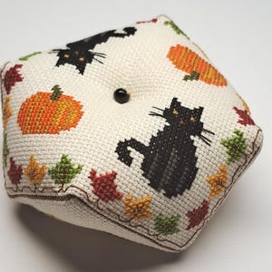 May include: A square, cream-coloured pin cushion with cross-stitched Halloween designs. It features black cats, orange pumpkins, and colourful autumn leaves. A black button is centered on the top. The edges are trimmed with brown stitching.