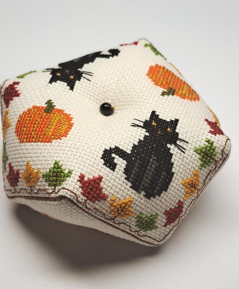 May include: A square, cream-coloured pin cushion with cross-stitched Halloween designs. It features black cats, orange pumpkins, and colourful autumn leaves. A black button is centered on the top. The edges are trimmed with brown stitching.