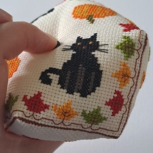May include: A cross-stitched cube with a black cat, pumpkins, and autumn leaves in various colours. The cube is made of off-white fabric with a brown border. A charming, handmade item, perfect for autumn or Halloween.