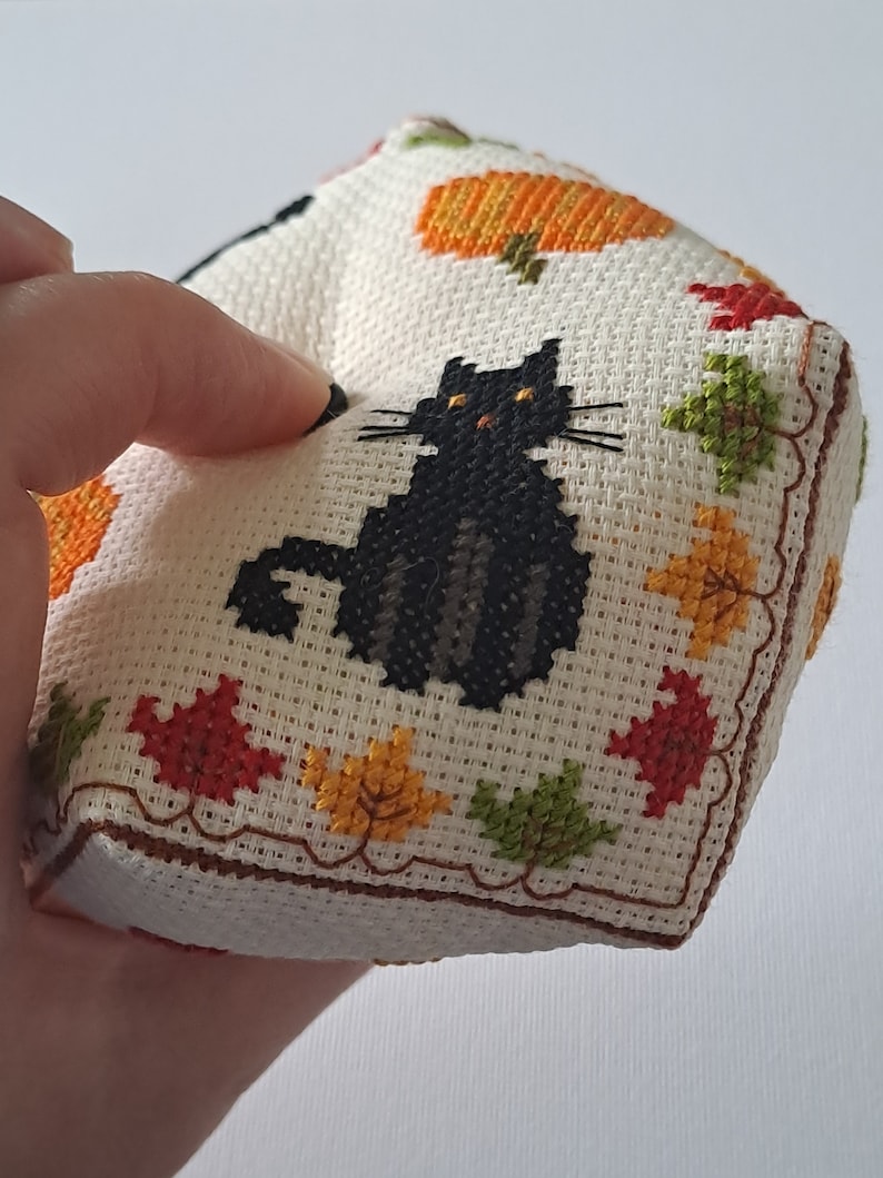 May include: A cross-stitched cube with a black cat, pumpkins, and autumn leaves in various colours. The cube is made of off-white fabric with a brown border. A charming, handmade item, perfect for autumn or Halloween.