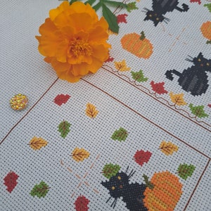 May include: Cross-stitch design with a black cat and orange pumpkin, surrounded by autumn leaves. The design is on a white background with a border of leaves and pumpkins. An orange flower and green leaves are in the foreground.