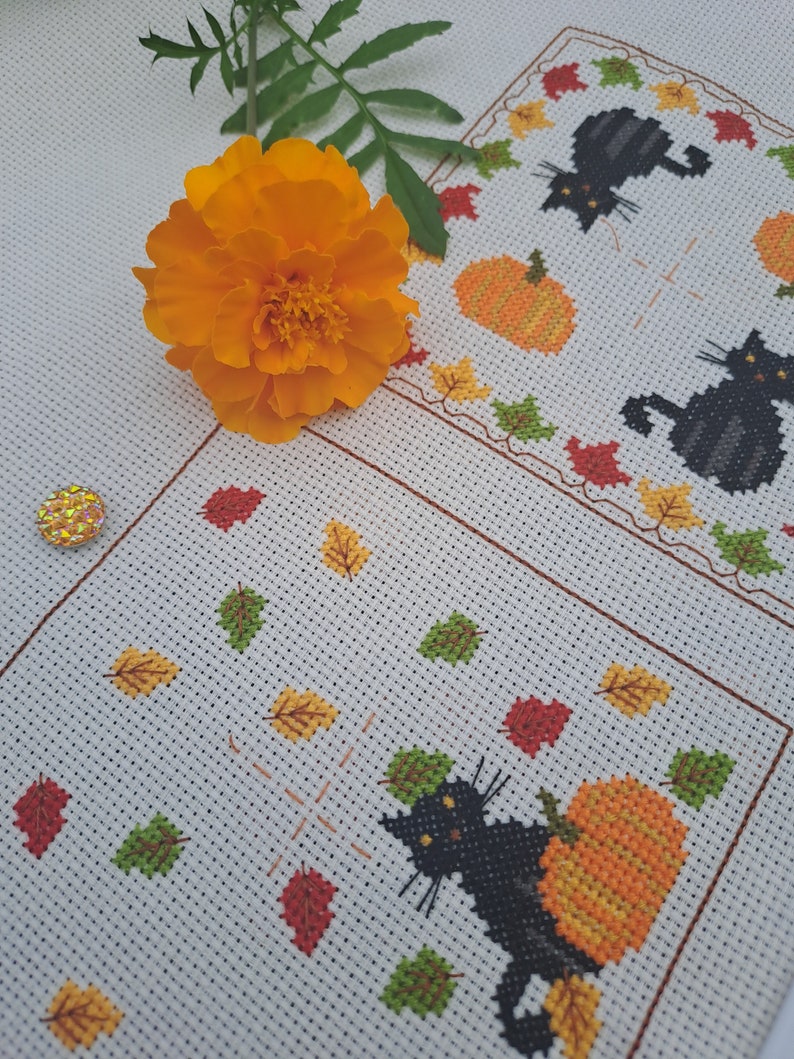 May include: Cross-stitch design with a black cat and orange pumpkin, surrounded by autumn leaves. The design is on a white background with a border of leaves and pumpkins. An orange flower and green leaves are in the foreground.