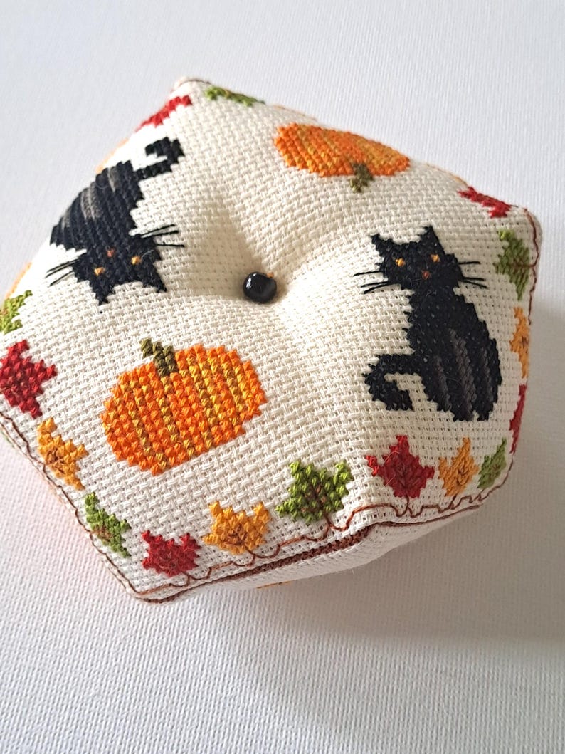 May include: A square, cream-coloured cross-stitch cushion featuring black cats and orange pumpkins. The cushion is decorated with autumn leaves in red, orange, and green. A black button is in the centre. The cushion is a decorative item.