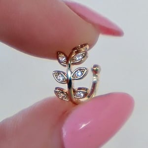 Puede incluir: Puño de oreja dorado con un diseño de hoja y acentos claros y brillantes. El puño se sostiene entre los dedos, mostrando sus delicados detalles y su elegante estilo. La joya está diseñada para ser usada en la oreja.