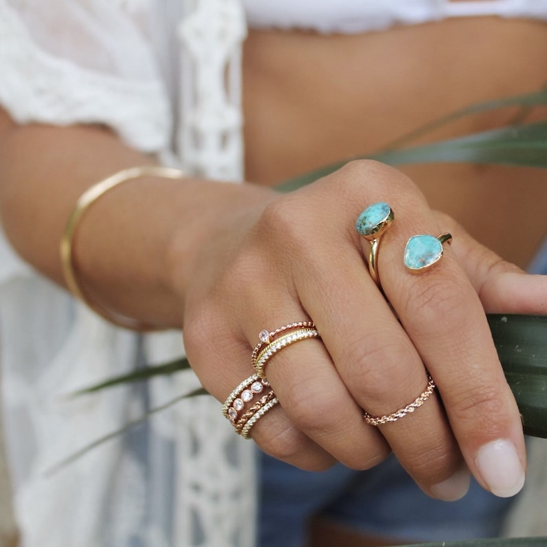May include: A woman's hand wearing a variety of gold rings, including a ring with two turquoise stones. The rings are stacked on her finger.