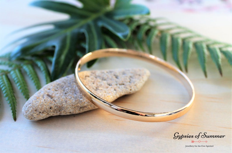 May include: A simple gold bangle bracelet, lying on a light brown surface with a small rock and green foliage in the background.