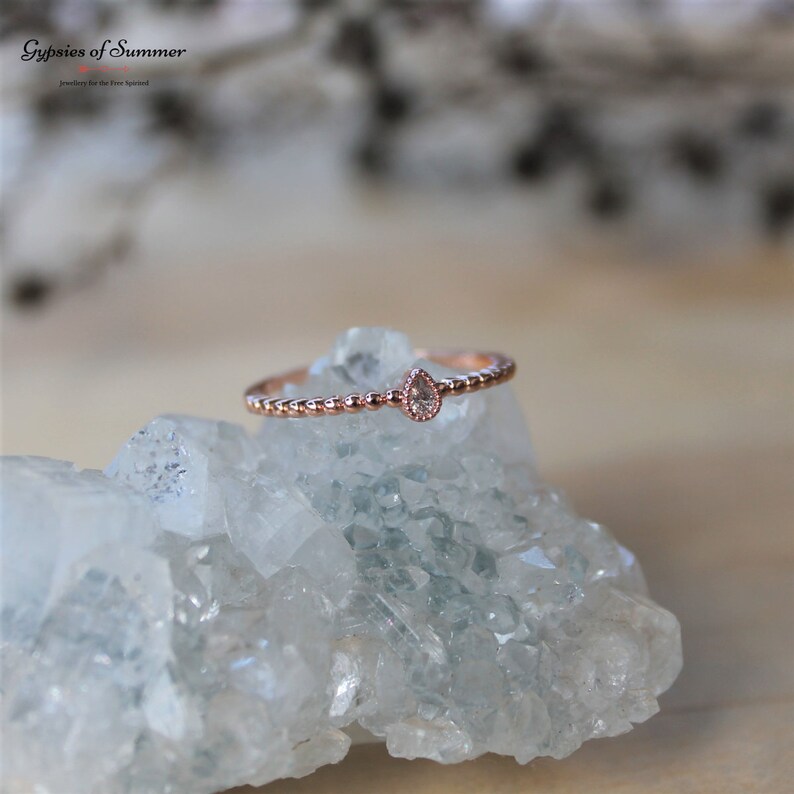 May include: A delicate rose gold ring with a small pear-shaped clear gemstone set in the center. The ring is beaded and sits on a white crystal background.