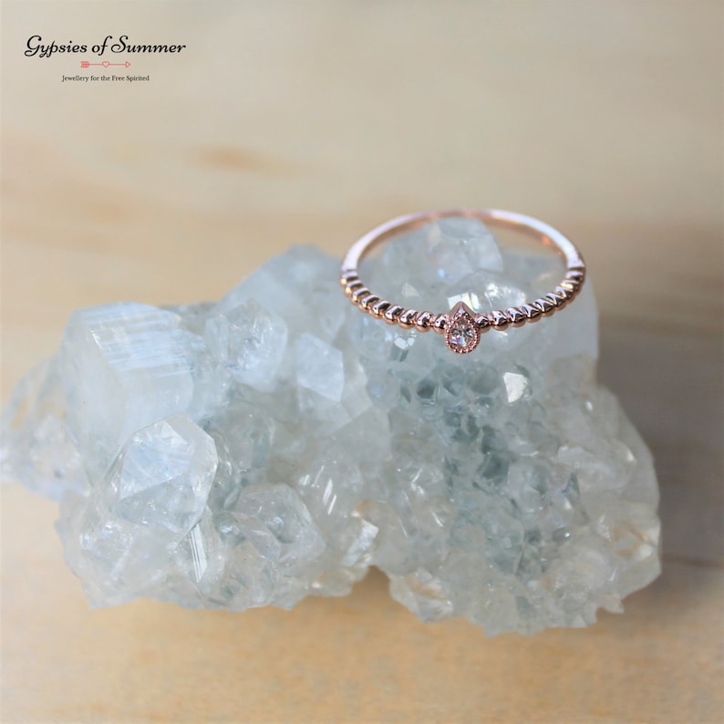 May include: A delicate rose gold ring with a small pear-shaped diamond set in the center. The ring is adorned with tiny beads along the band.