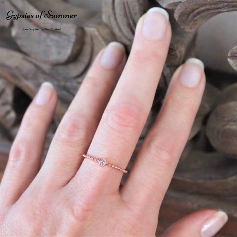 May include: A delicate rose gold ring with a small, clear gemstone set in the center. The ring is on a finger, with the background out of focus.