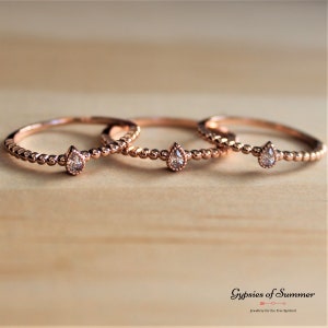 May include: Three delicate rose gold rings with a pear-shaped clear gemstone on each. The rings are stacked on a wooden surface.