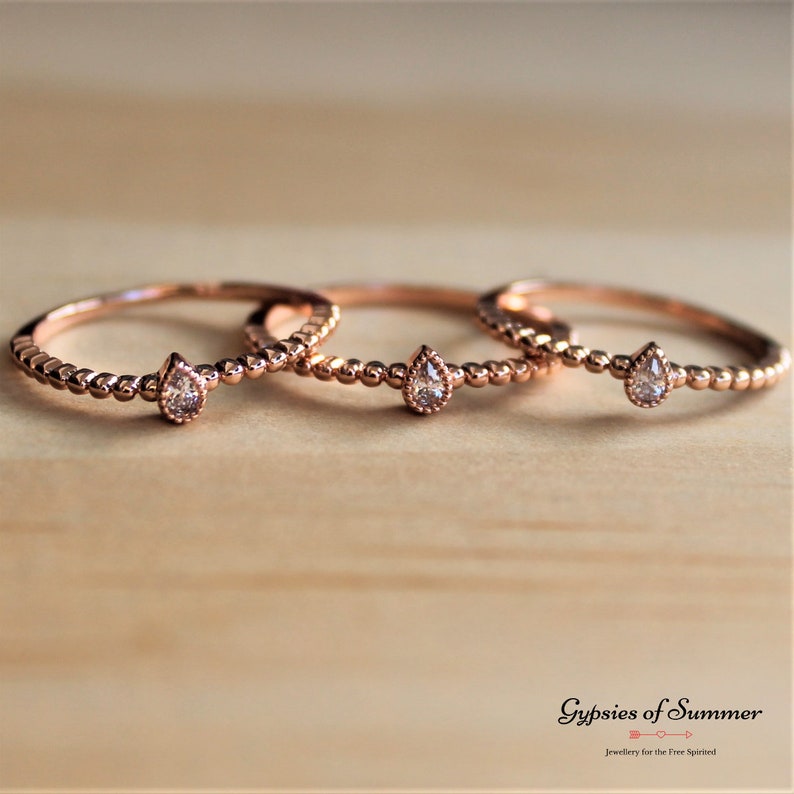May include: Three delicate rose gold rings with a pear-shaped clear gemstone on each. The rings are stacked on a wooden surface.