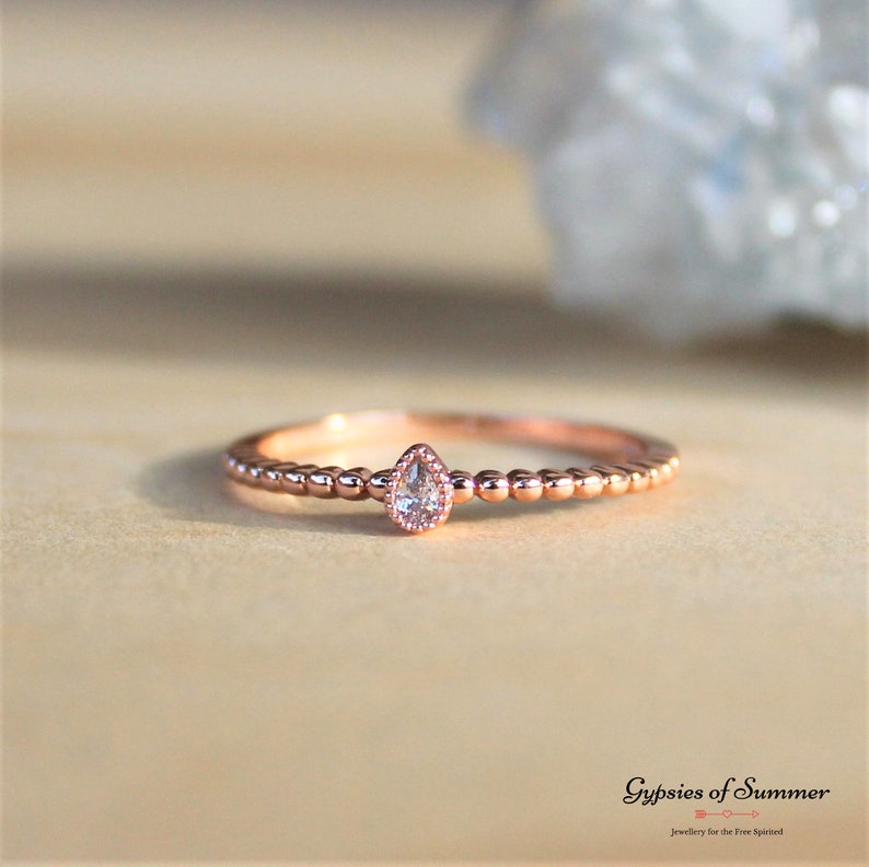 May include: A delicate rose gold ring with a pear-shaped clear gemstone set in the center. The band is adorned with small beads.