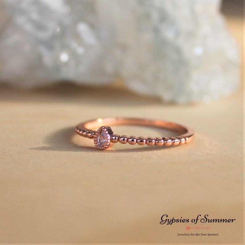 May include: A delicate rose gold ring with a single clear pear-shaped stone set in the center. The band is adorned with small beads.