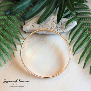 May include: A simple gold bangle bracelet, shown on a wooden surface with green foliage.