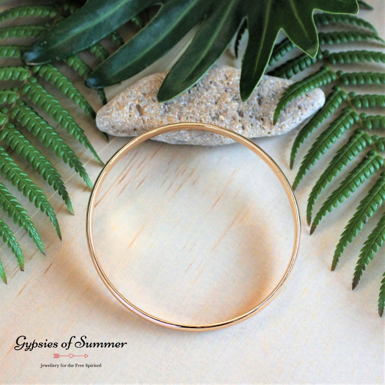 May include: A simple gold bangle bracelet, shown on a wooden surface with green foliage.