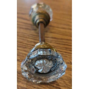 May include: A vintage glass doorknob with a faceted design. The knob has a clear, sparkling appearance and is attached to a brass-colored metal base and a brown metal rod. The doorknob is likely for a cabinet or a door.