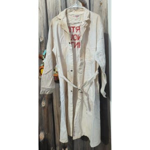 May include: A long-sleeved, off-white duster coat featuring a pointed collar, a button-down front, and a matching tie belt at the waist. The chest area displays red text that reads "ART" and "YOU" stacked vertically.